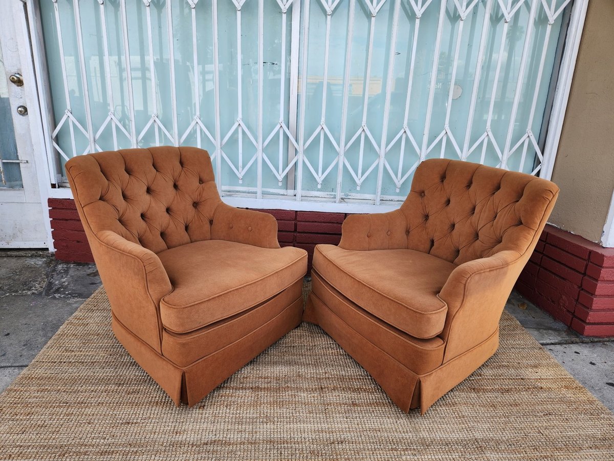 Two tan upholstered armchairs with tufted backs facing each other on a beige rug, with white curtains and brown storage in background.