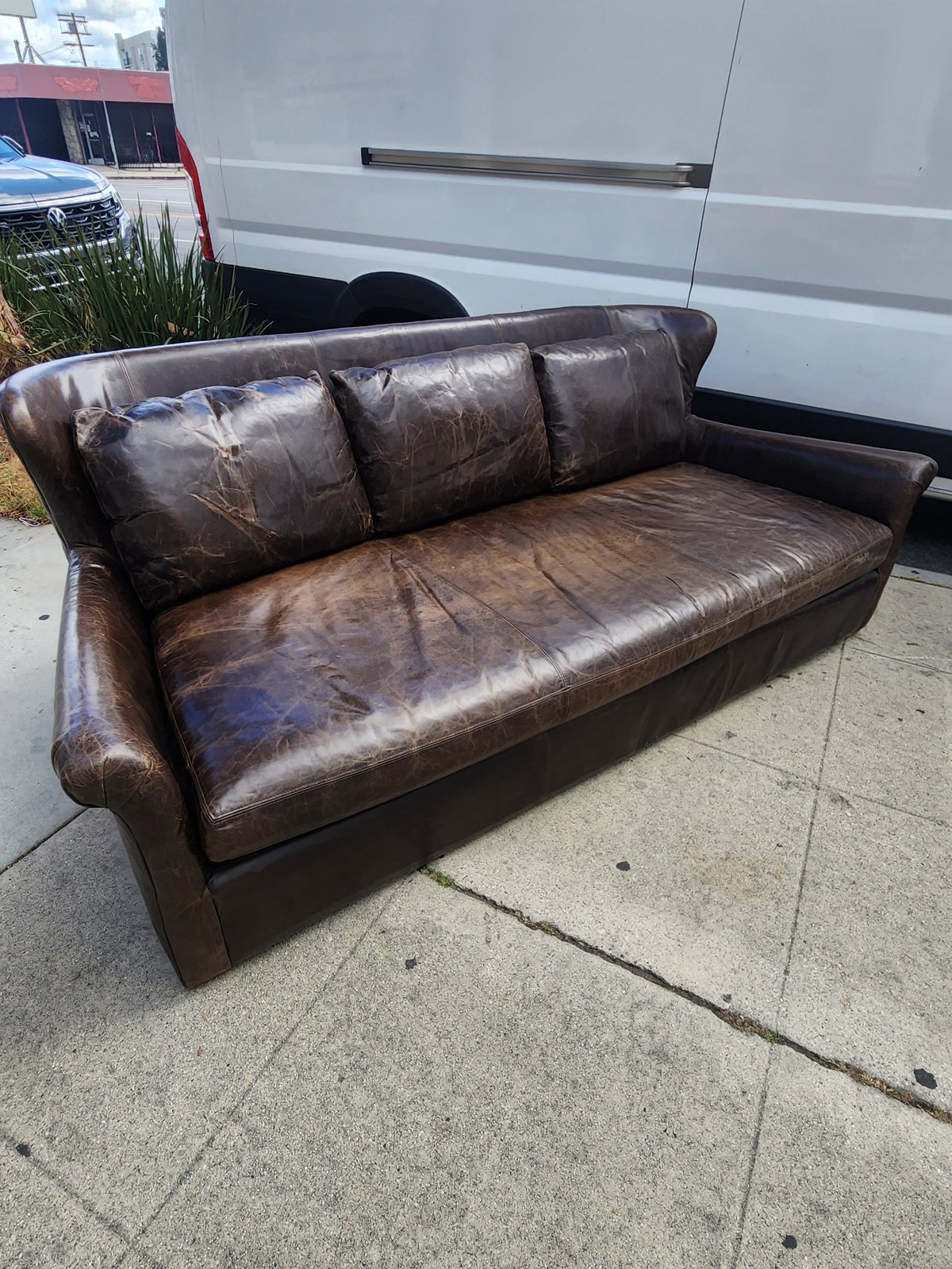 Worn brown leather sofa sitting on concrete sidewalk in front of white delivery van