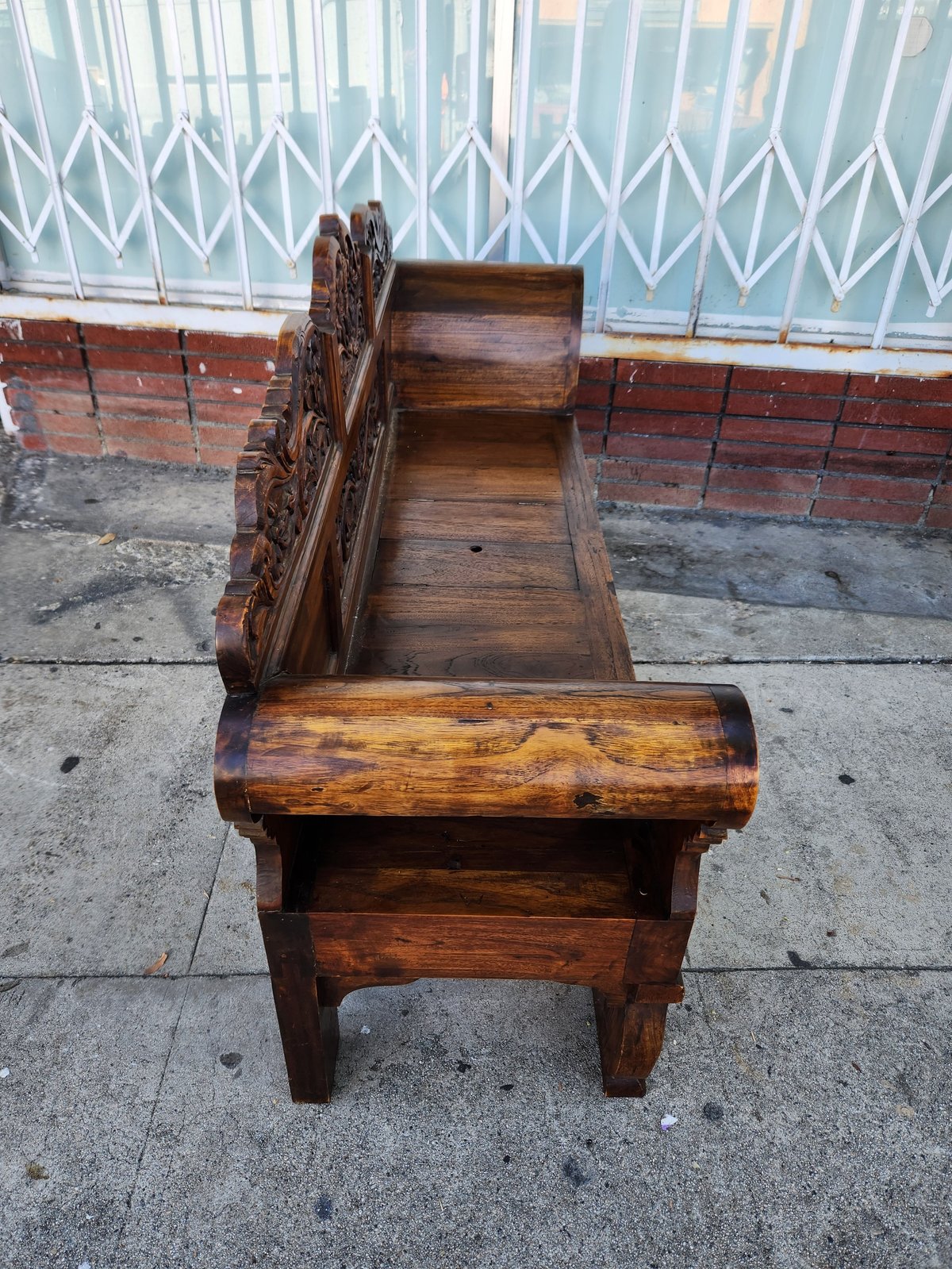 Antique carved wooden bench with ornate arm and storage compartments on concrete pavement in front of metal gate
