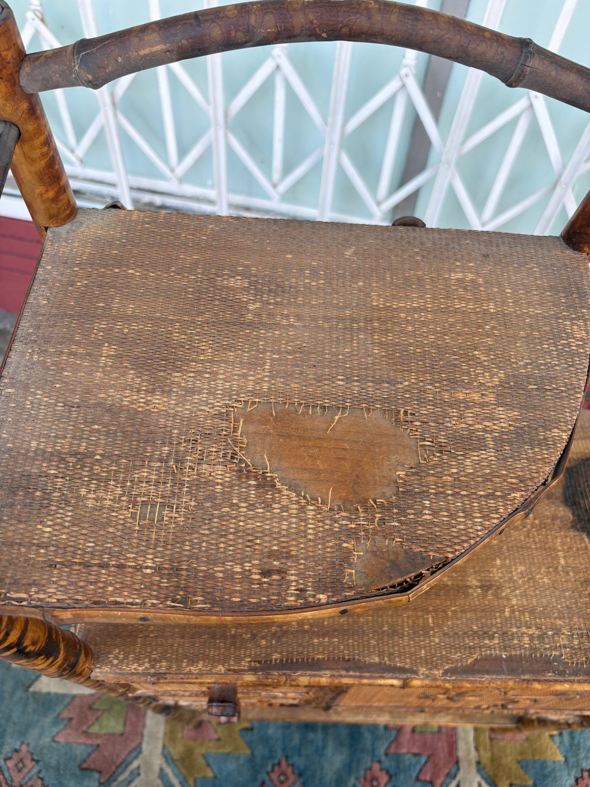 Worn wooden chair with woven wicker seat showing stains and wear, against geometric patterned wallpaper background
