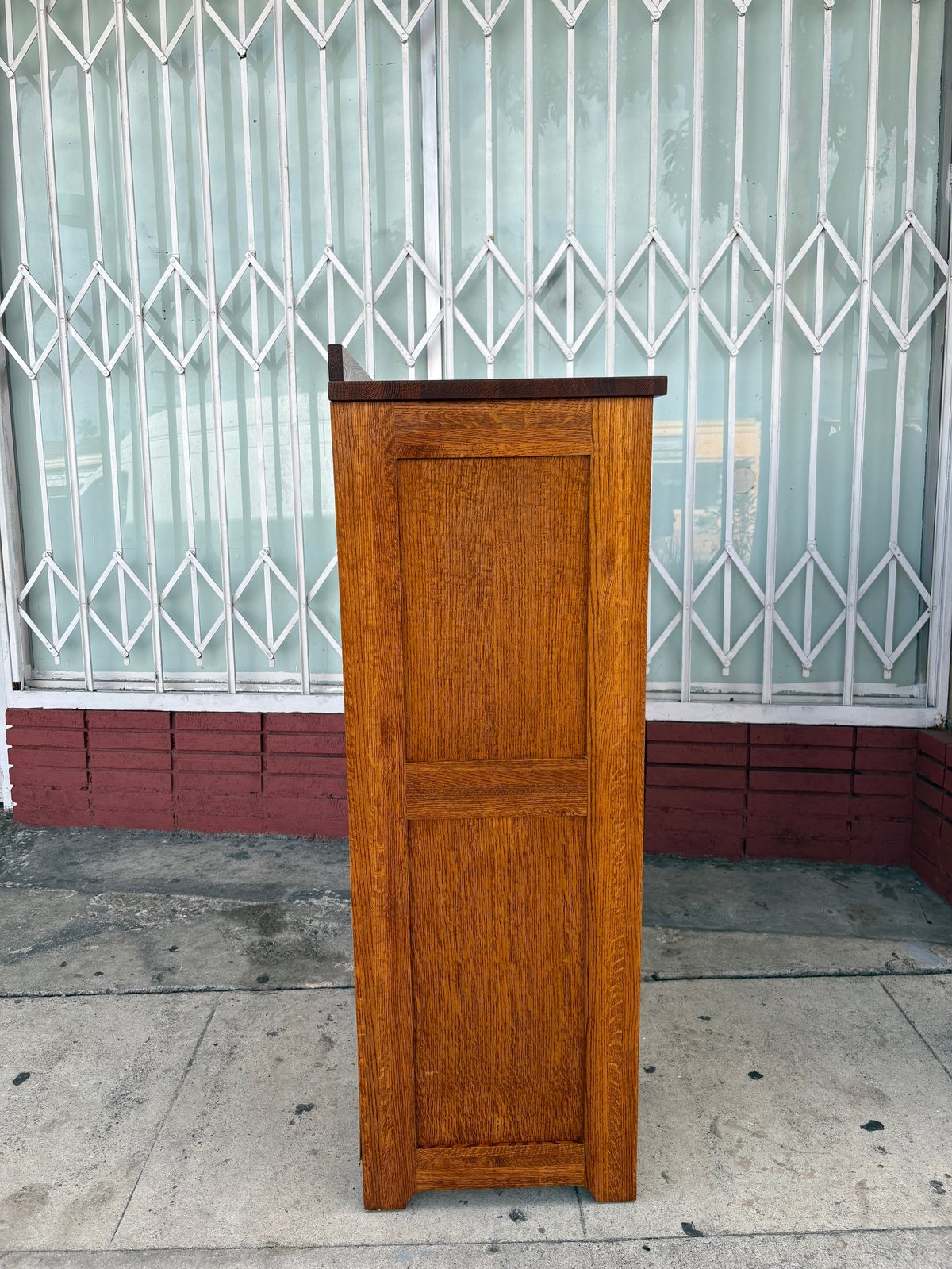 Wooden cabinet with two panel doors standing on pavement in front of metal security gate and red wall