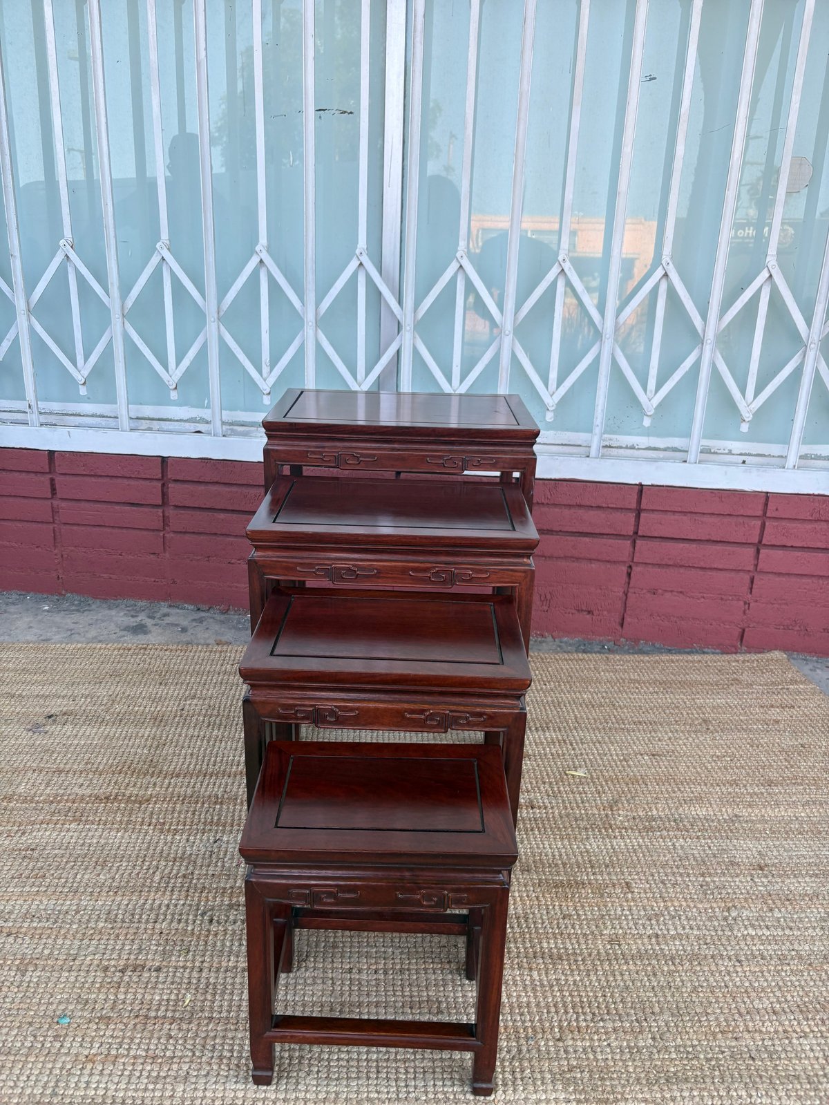 Stacked set of four wooden nesting tables with dark cherry finish against a white lattice gate and red brick wall