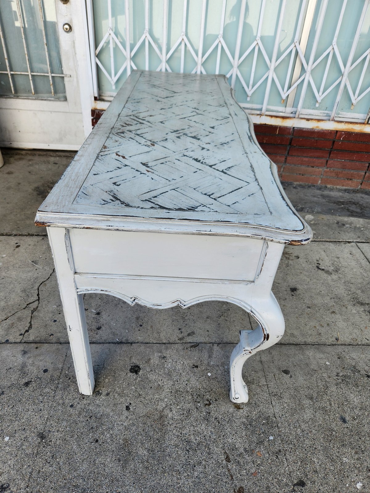 Whitewashed wooden console table with curved legs on concrete patio, featuring a distressed blue-gray marble-effect top