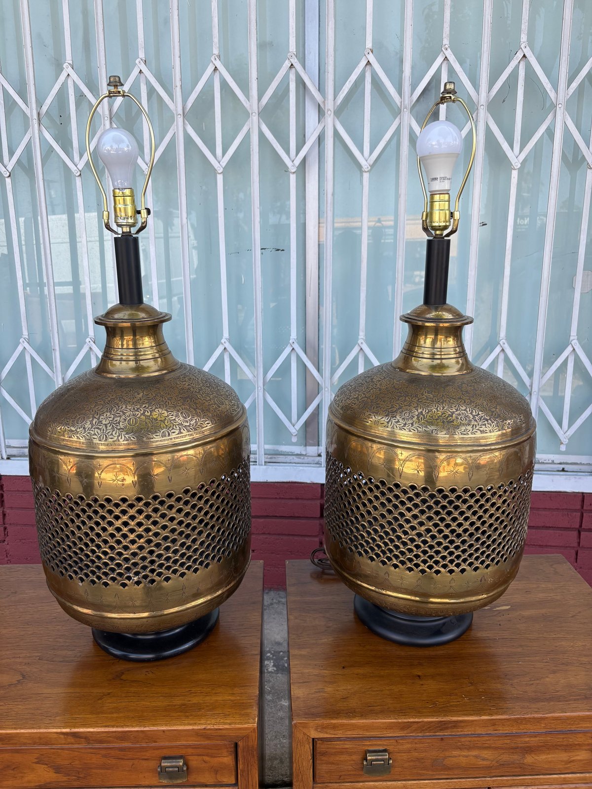 Pair of brass cylindrical table lamps with mesh detailing on wooden surface against patterned curtain backdrop