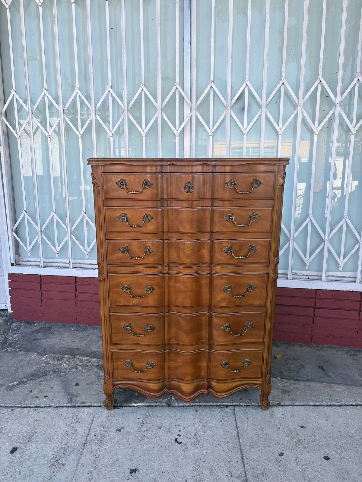 Antique wooden dresser with multiple drawers and brass handles displayed on a sidewalk against a white security gate.