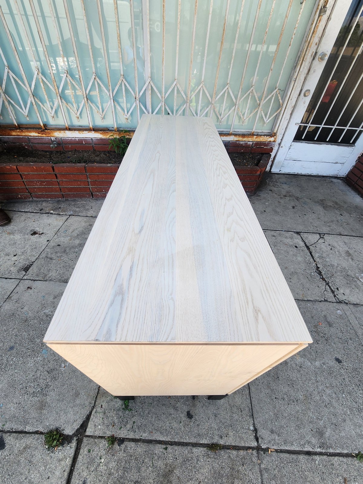 Long white wooden table on wheels outdoors, with corrugated metal gate and door visible in background