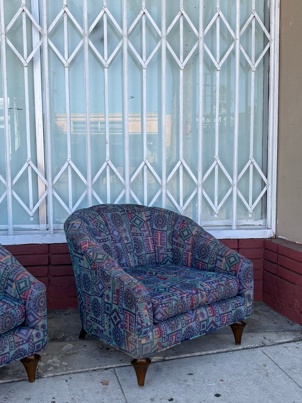 Patterned upholstered armchair outside storefront with white metal accordion security gate and red brick base