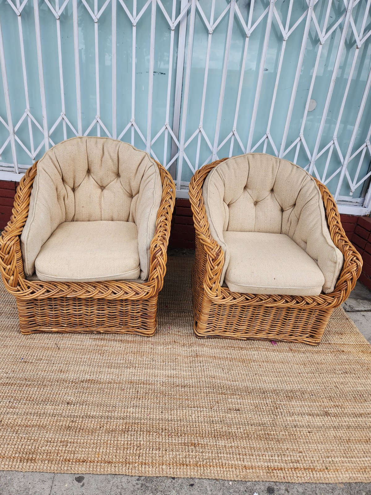 Two wicker armchairs with cream cushions on a beige rug in front of a white patterned metal gate