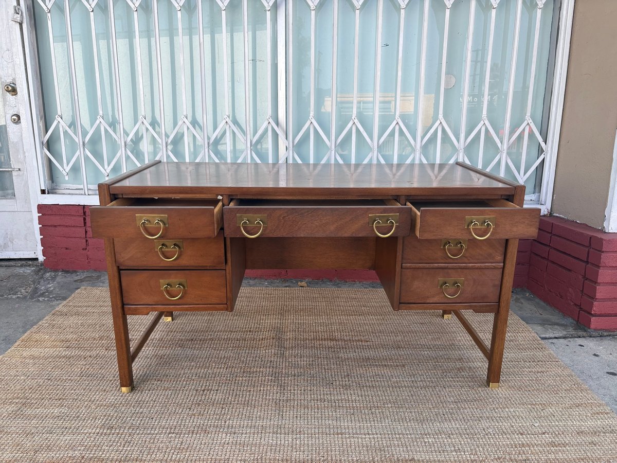 Wooden mid-century desk with six drawers and brass handles on a tan area rug, set against white security gates.