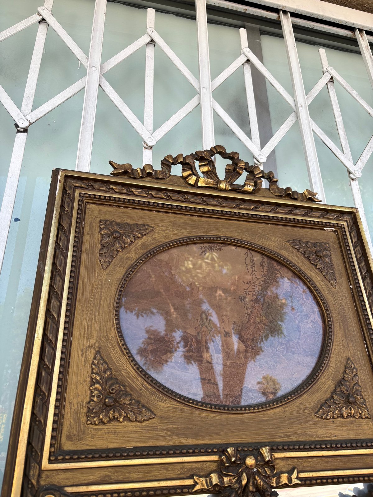 Ornate gold-framed portrait painting with oval centerpiece and decorative corner flourishes, mounted on white geometric wall paneling