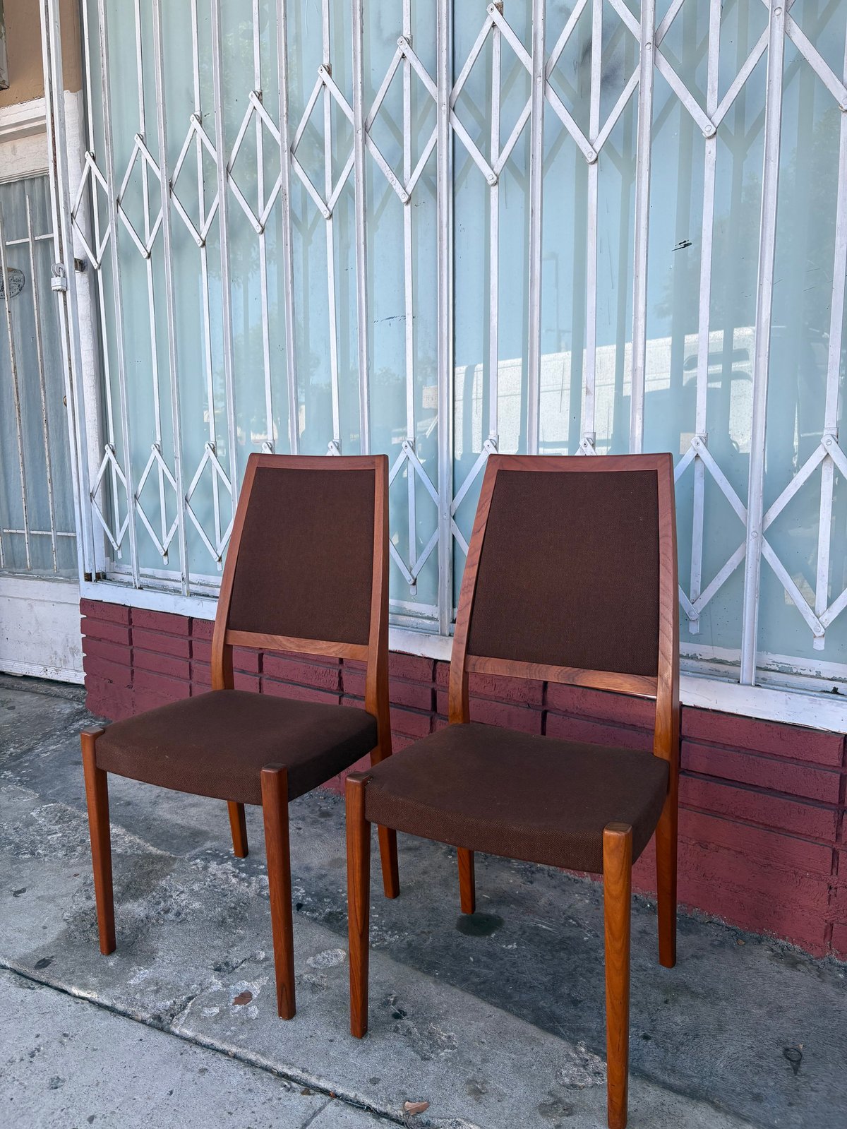 Two brown upholstered dining chairs with wooden legs positioned on a sidewalk in front of a metal security gate