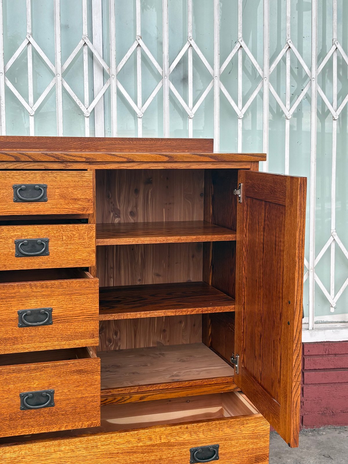 Vintage wooden cabinet with open door and shelves, featuring drawers with metal handles, against a white latticed window background