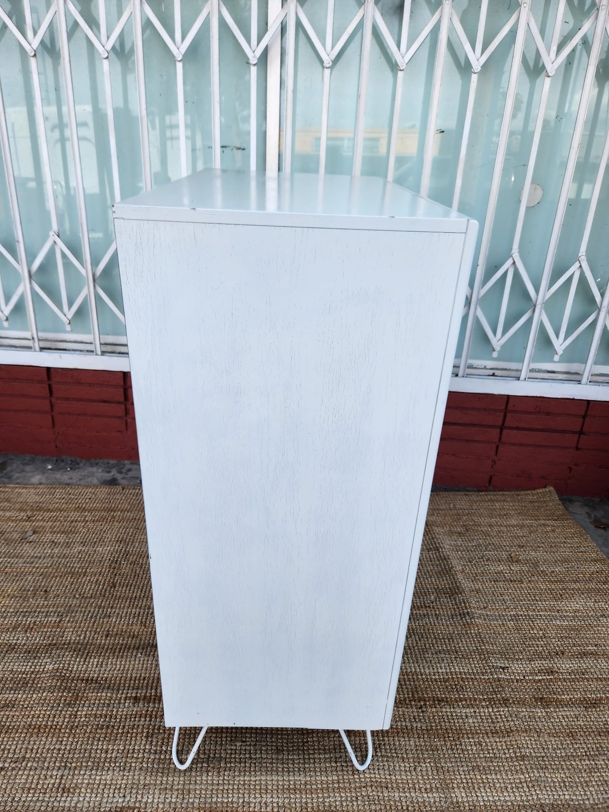 White modern storage cabinet with hairpin legs on beige carpet, metal gate visible behind