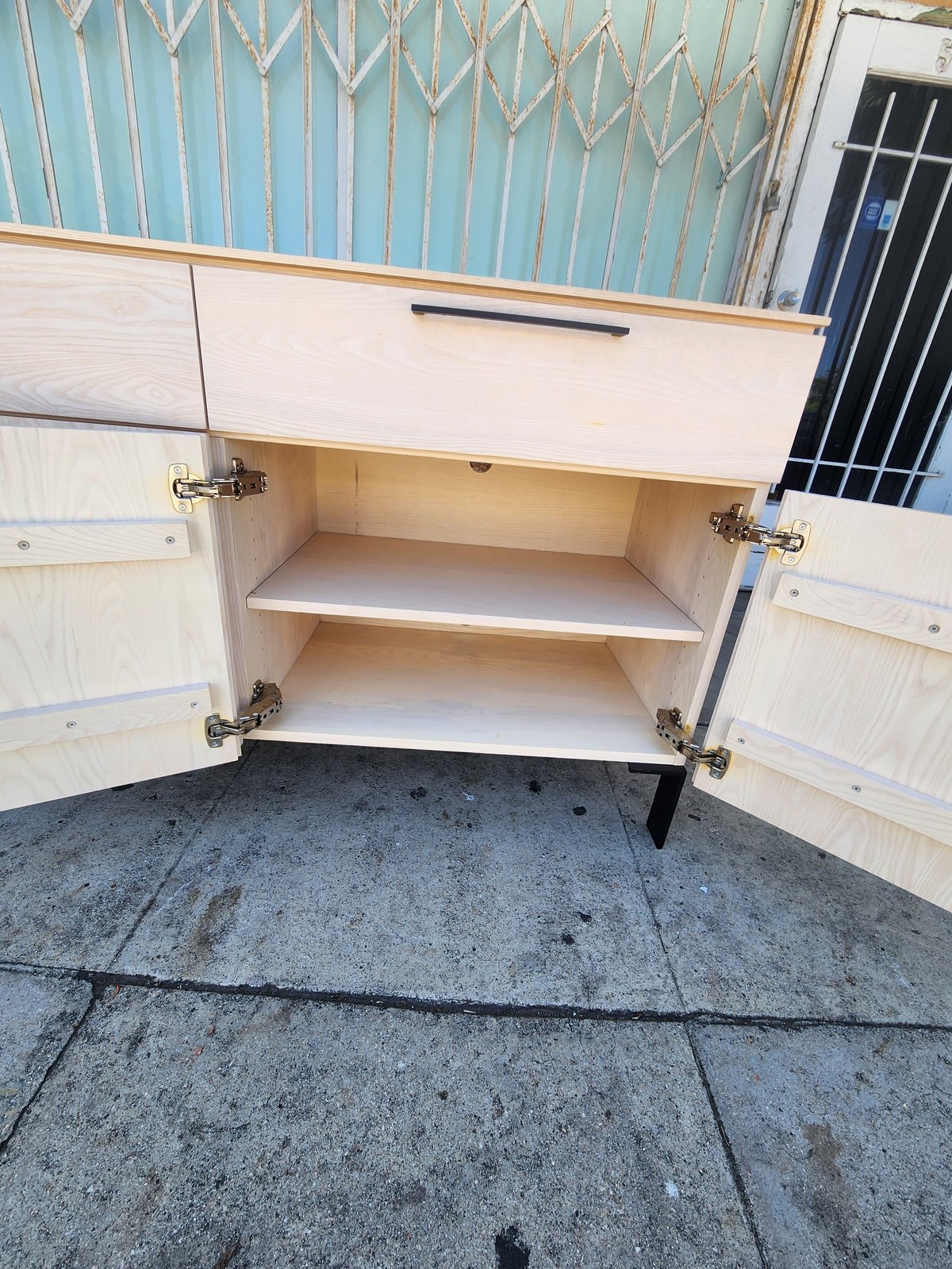 Open wooden cabinet with two shelves and white doors on concrete floor against blue metal siding wall