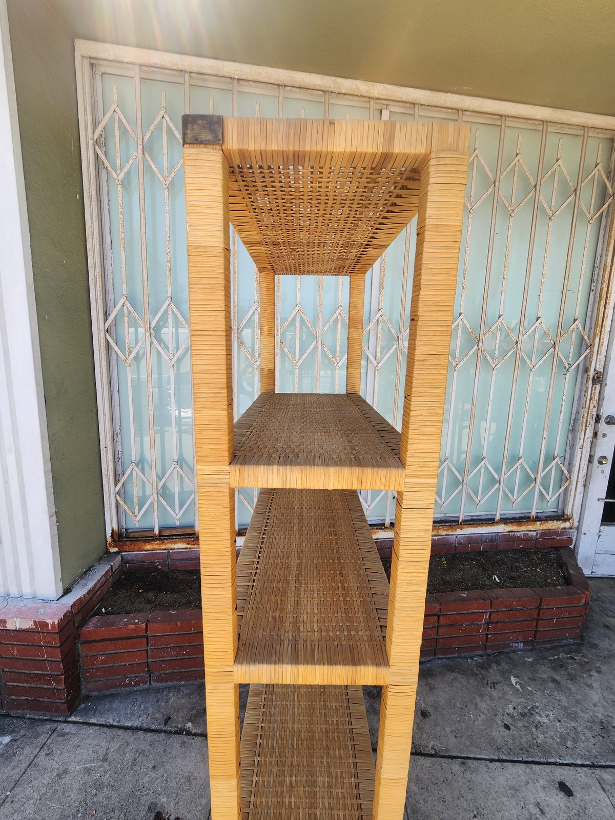Wooden shelving unit with woven rattan shelves positioned in front of blue patterned glass doors and brick base on pavement