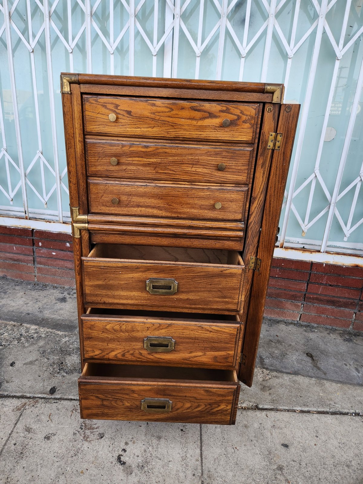 Wooden dresser with multiple drawers and brass hardware standing on concrete against white metal gate