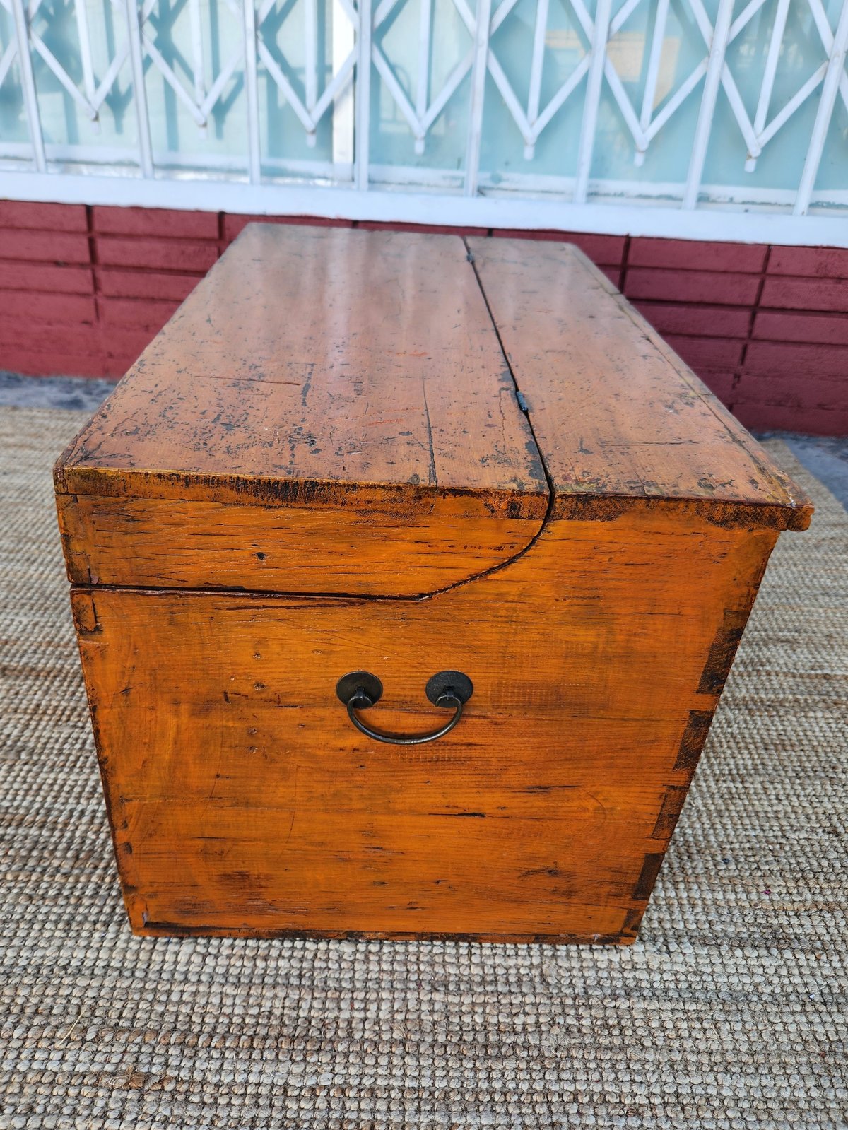 Vintage wooden trunk with metal handles and rope, placed on pavement against a red and white building background