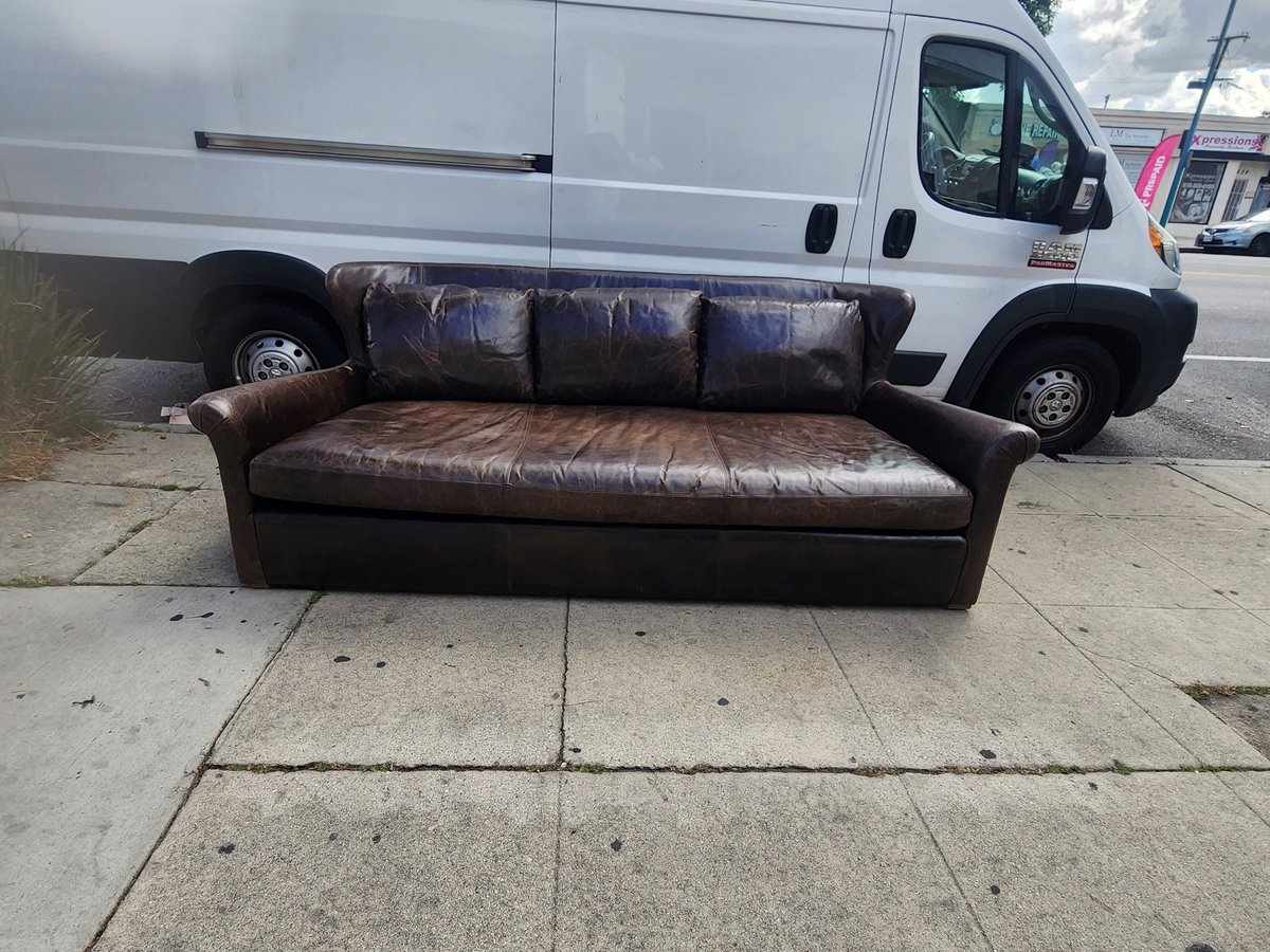 Worn brown leather sofa sitting on concrete sidewalk with white van parked behind it