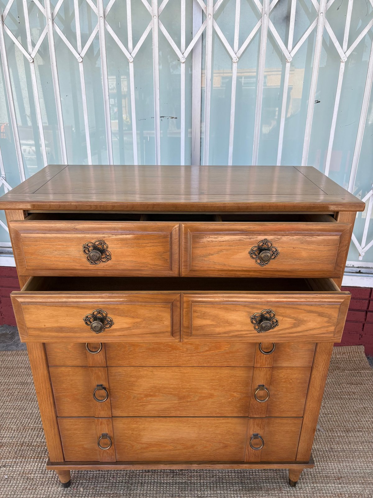 Wooden dresser with four drawers and decorative hardware, displayed indoors against white curtained windows.
