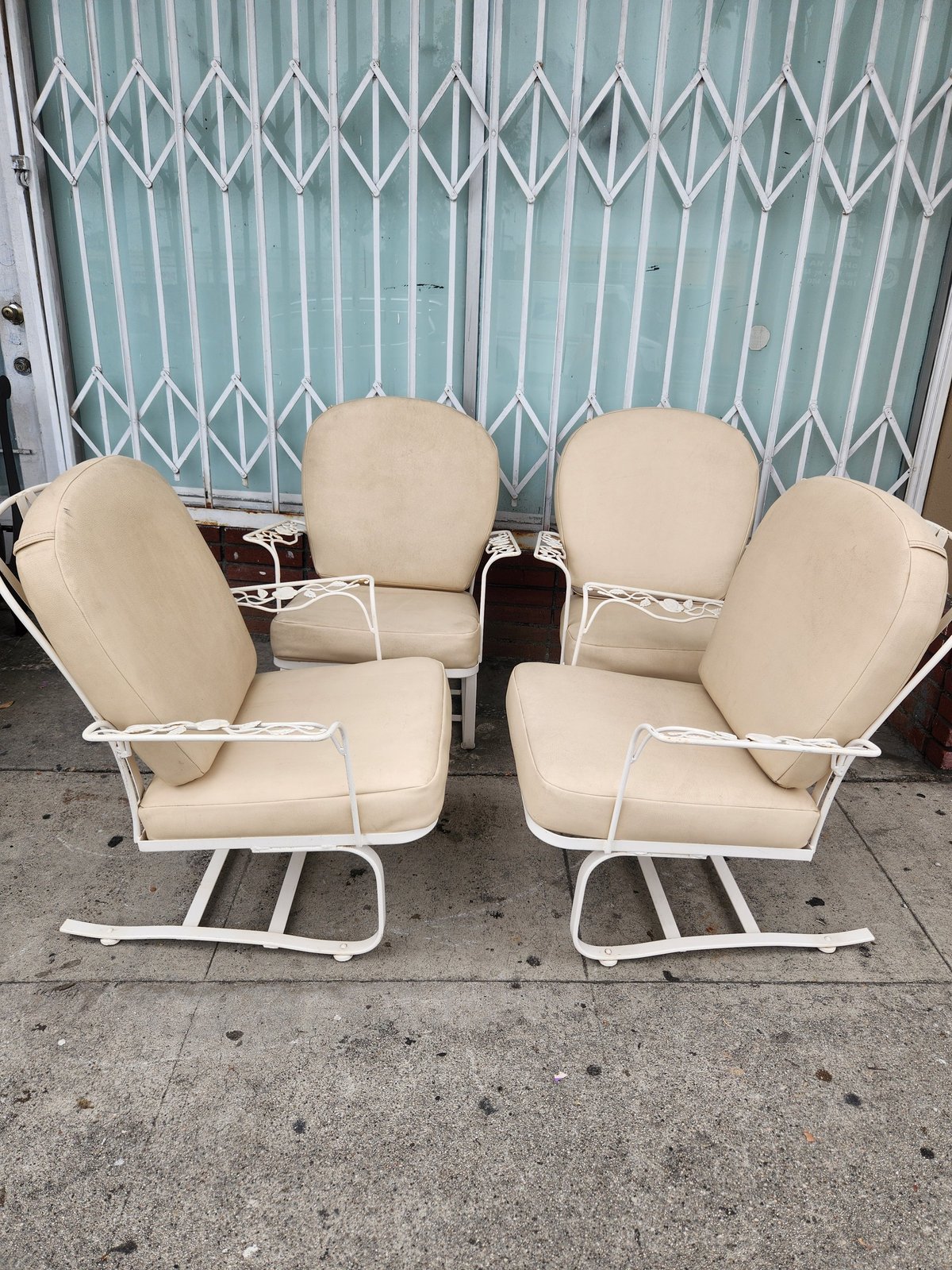 Four cream-colored metal outdoor chairs with cushions arranged in a circle against a turquoise metal corrugated gate