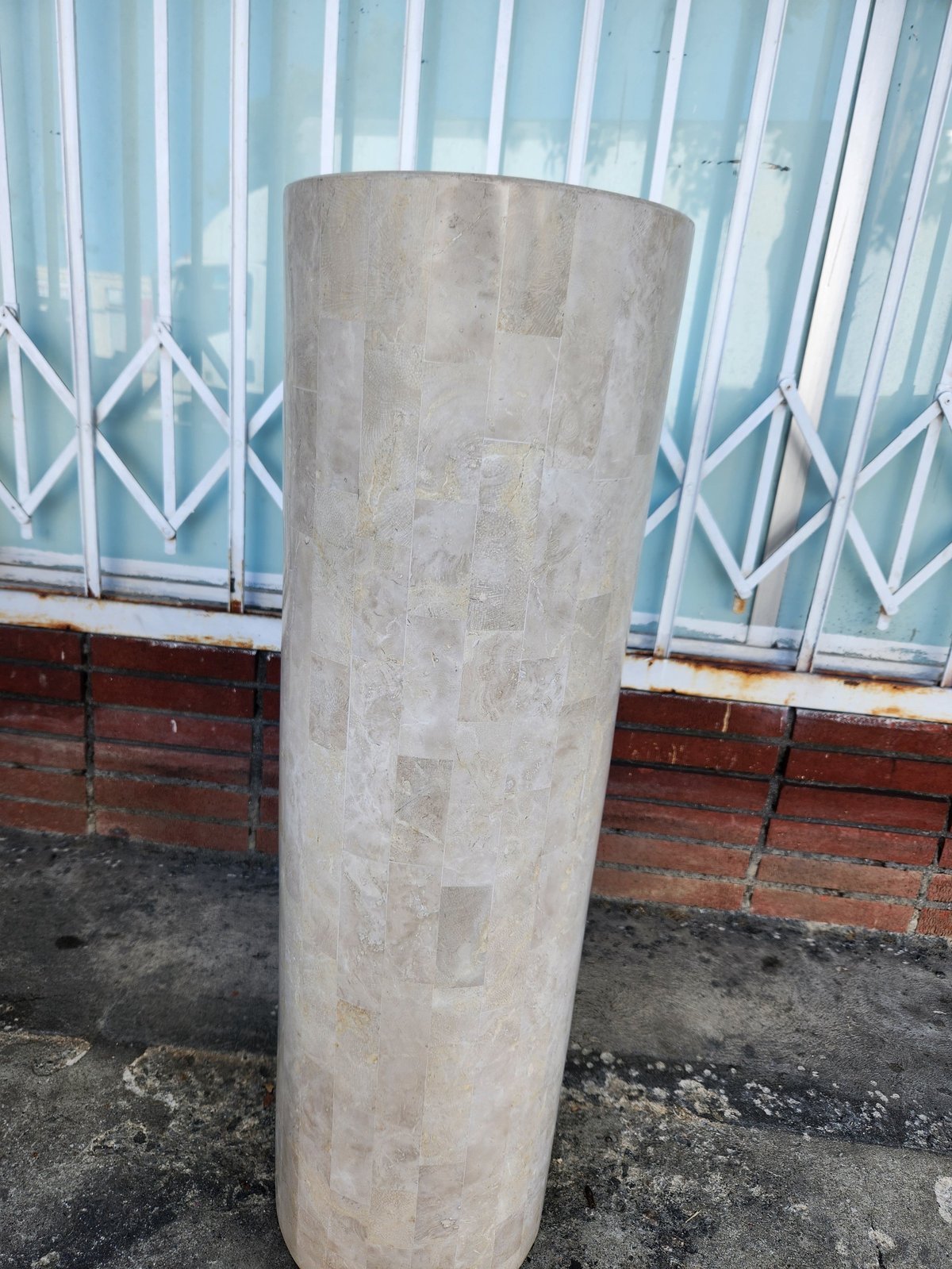 Concrete cylindrical column standing on ground in front of corrugated metal fence and brick wall