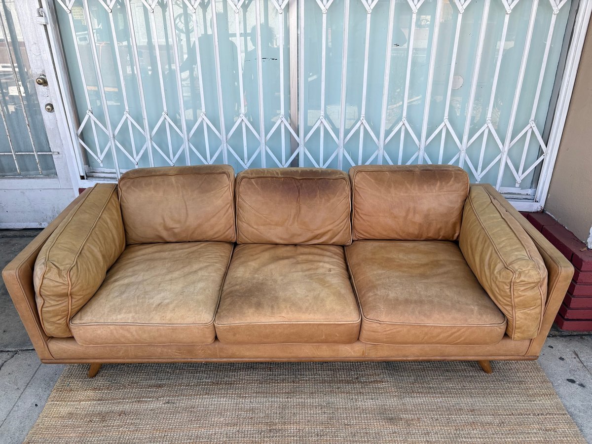 Tan leather three-seat sofa with rolled arms and wooden legs displayed on a sidewalk with white security gate in background