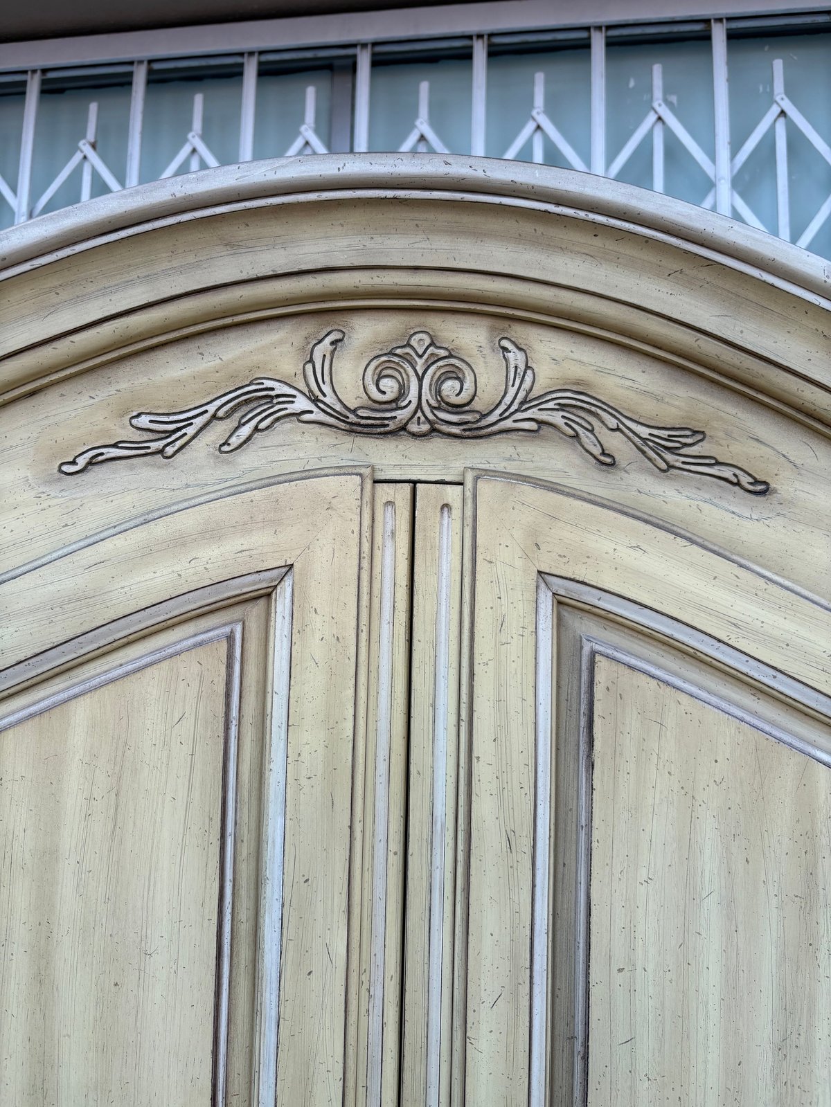 Ornate pale wooden double doors with decorative carved scrollwork above the frame and geometric transom window detailing