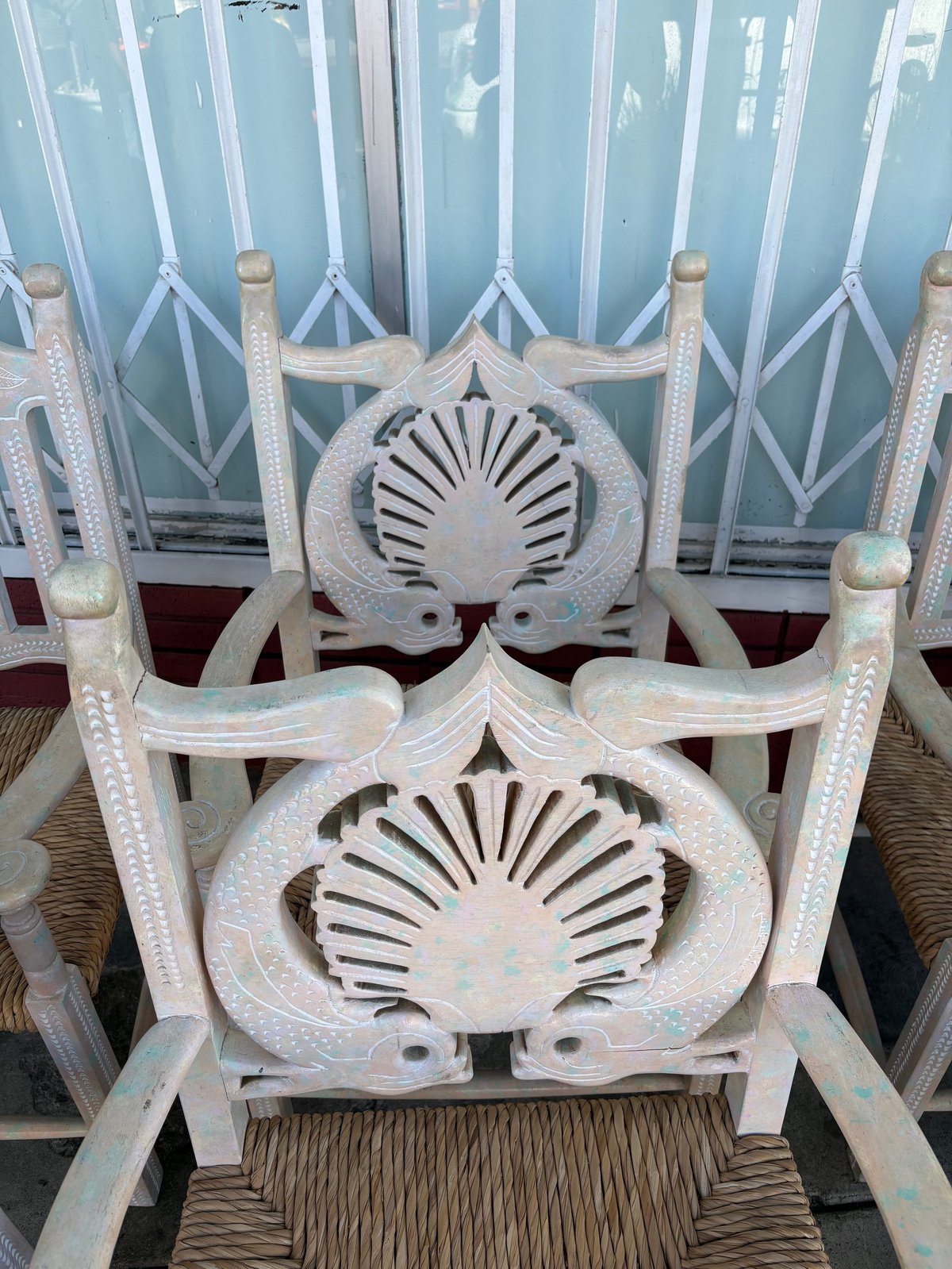 White ornate wooden chair with shell-shaped carved backrest, positioned outdoors with similar chairs visible in background