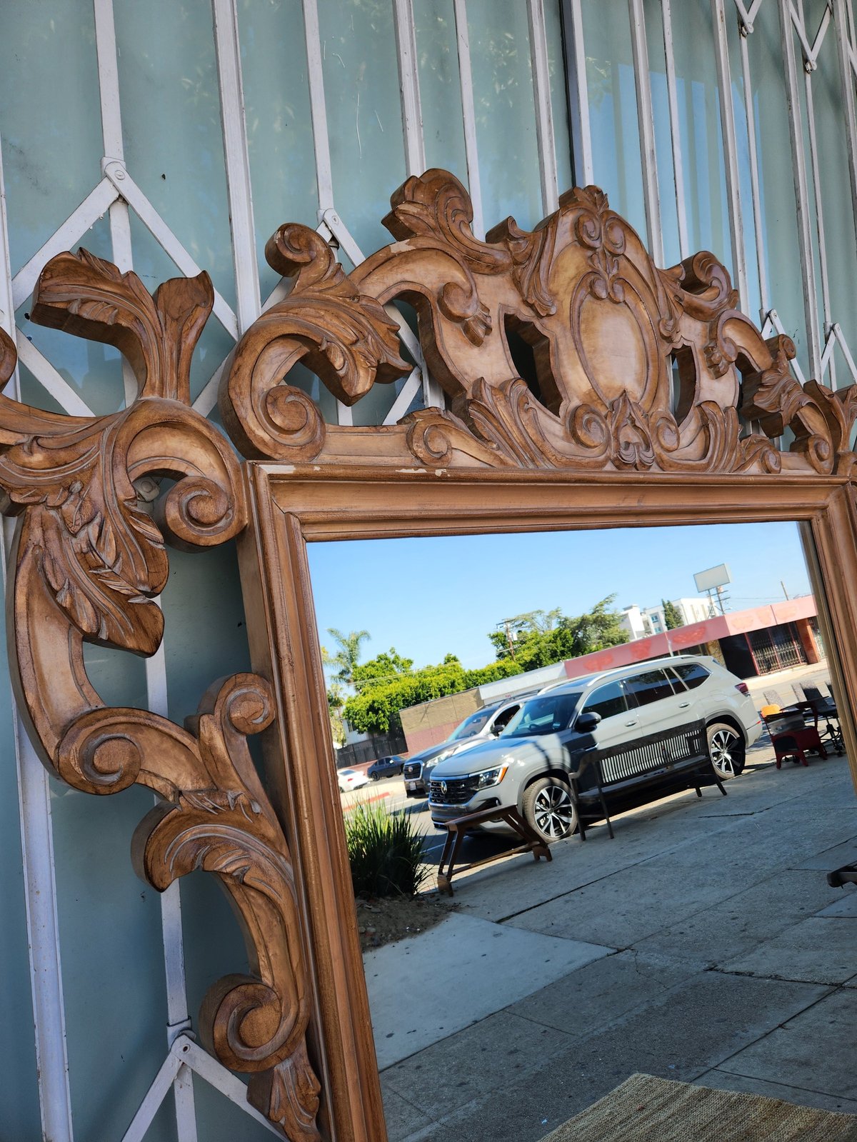 Ornate carved wooden mirror frame with decorative scrollwork reflecting a parking lot with a vehicle and buildings