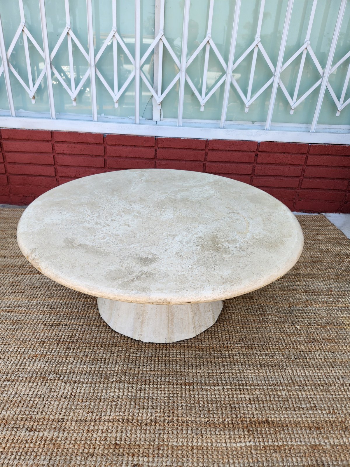 Cream-colored round marble pedestal table on beige carpet with red brick wall and white decorative gate behind it