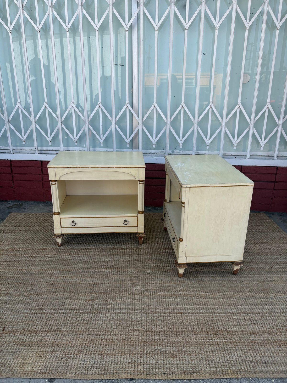 Two cream-colored wooden nightstands with drawers displayed on pavement in front of a folding metal gate and red brick wall