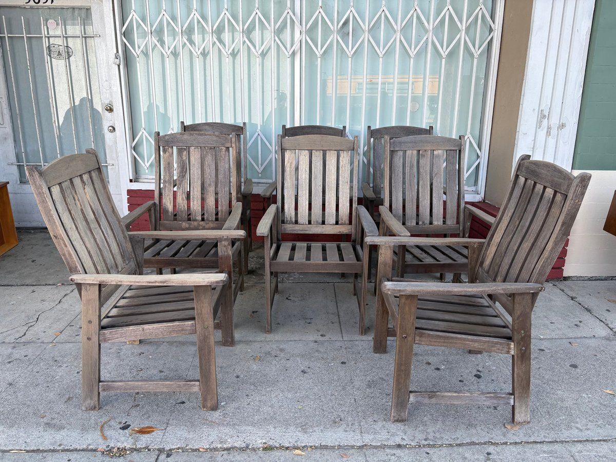 Five wooden outdoor chairs arranged on a concrete patio in front of a house with venetian blinds