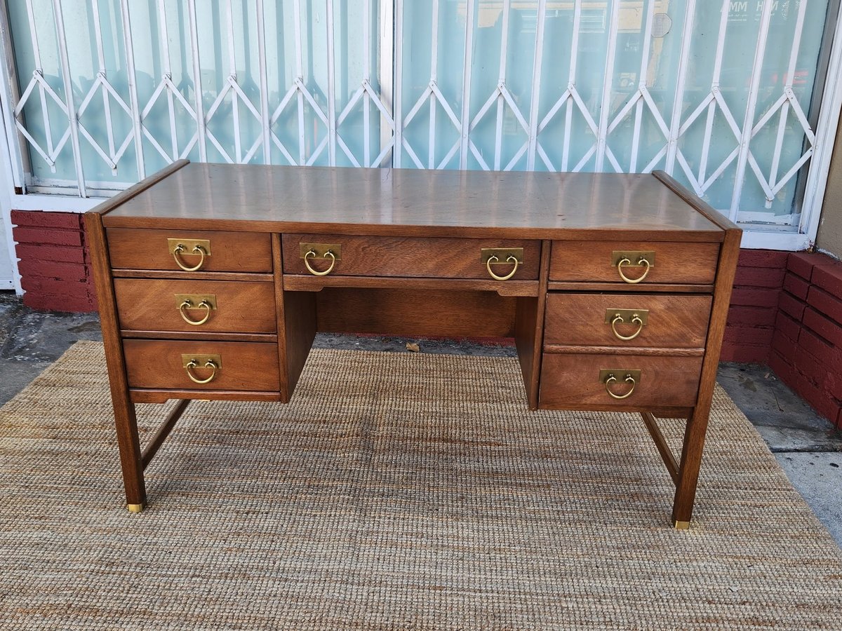 Vintage wooden desk with six drawers and brass handles, placed on a beige rug in front of a window with white curtains