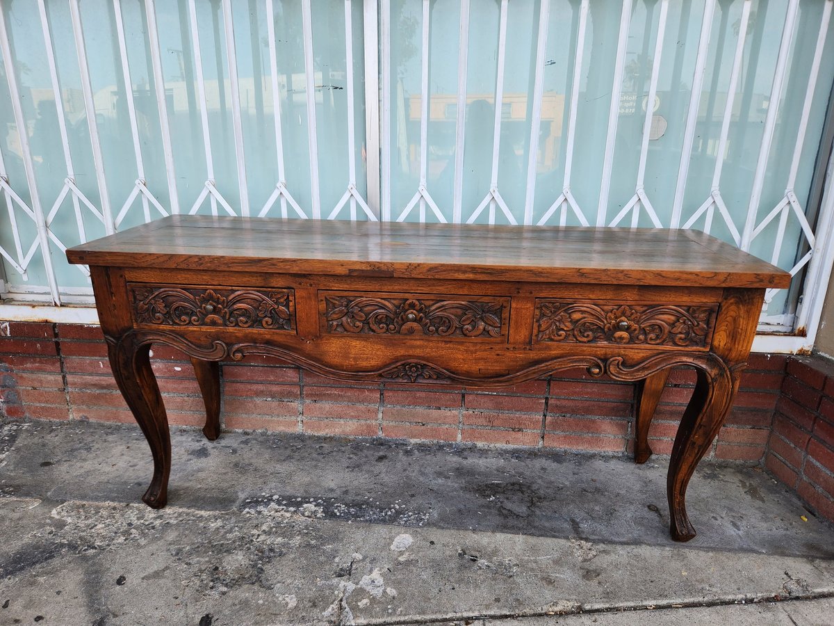 Wooden console table with ornate carved drawers and curved cabriole legs against white curtains