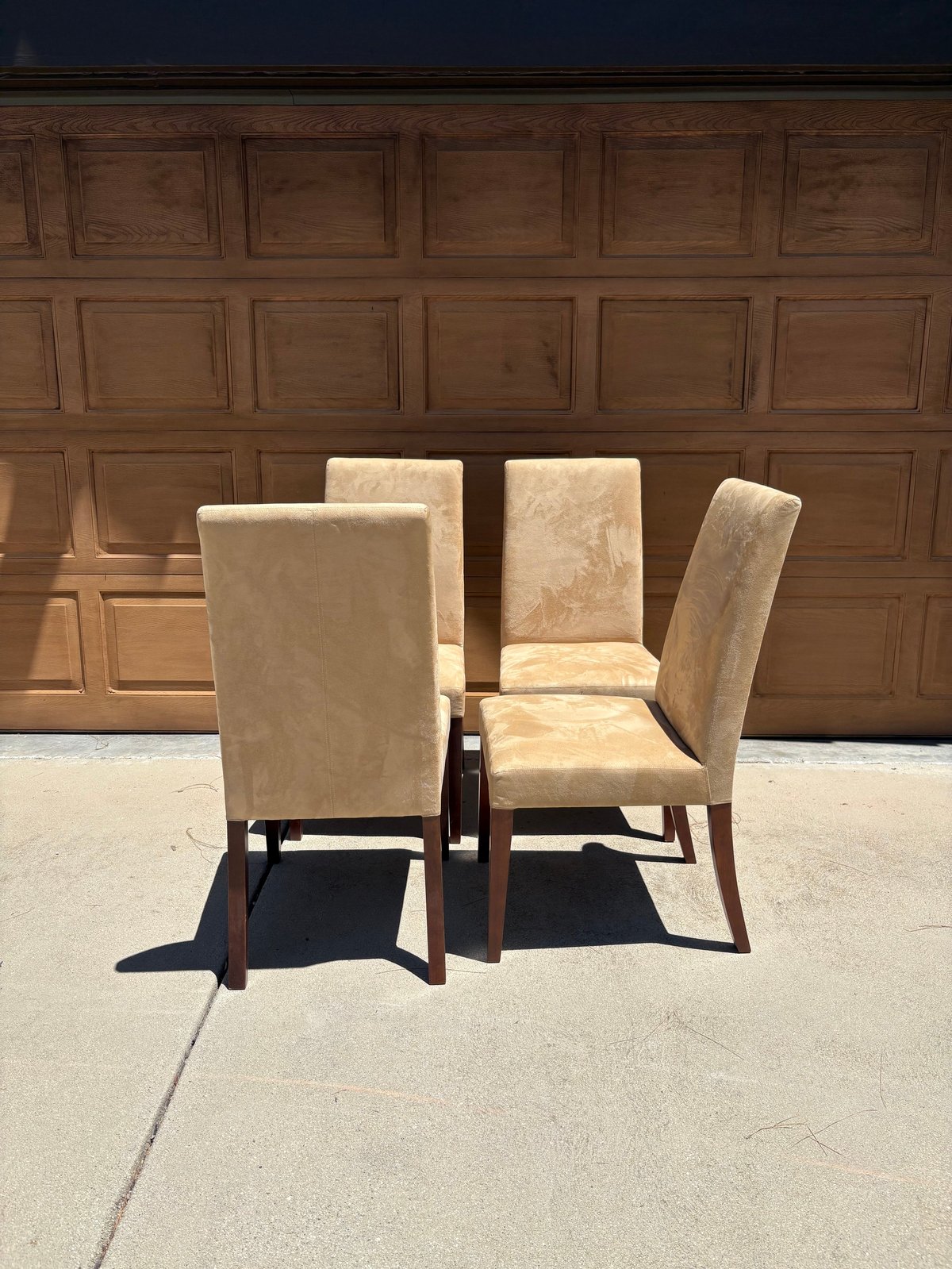 Four upholstered dining chairs with beige fabric and wooden legs arranged on concrete driveway in front of brown wooden garage door
