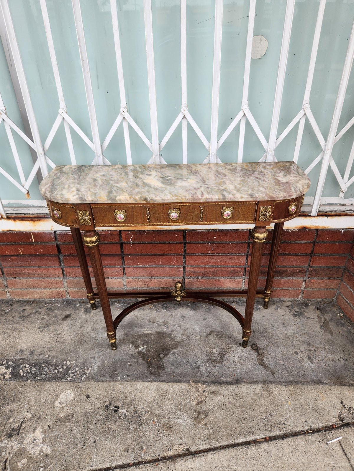 Antique wooden console table with marble top and brass legs, positioned against a white painted fence on a brick wall