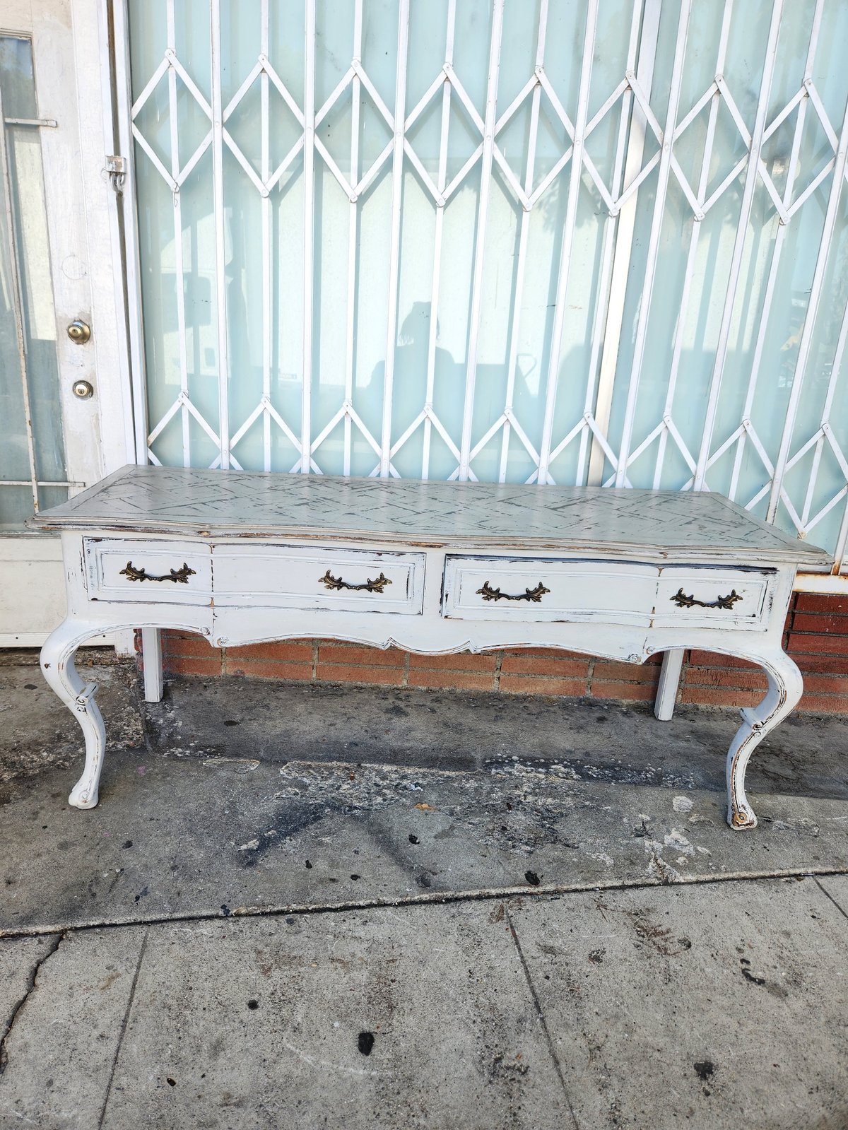 White vintage console table with curved legs and three drawers positioned on concrete ground in front of metal security gate