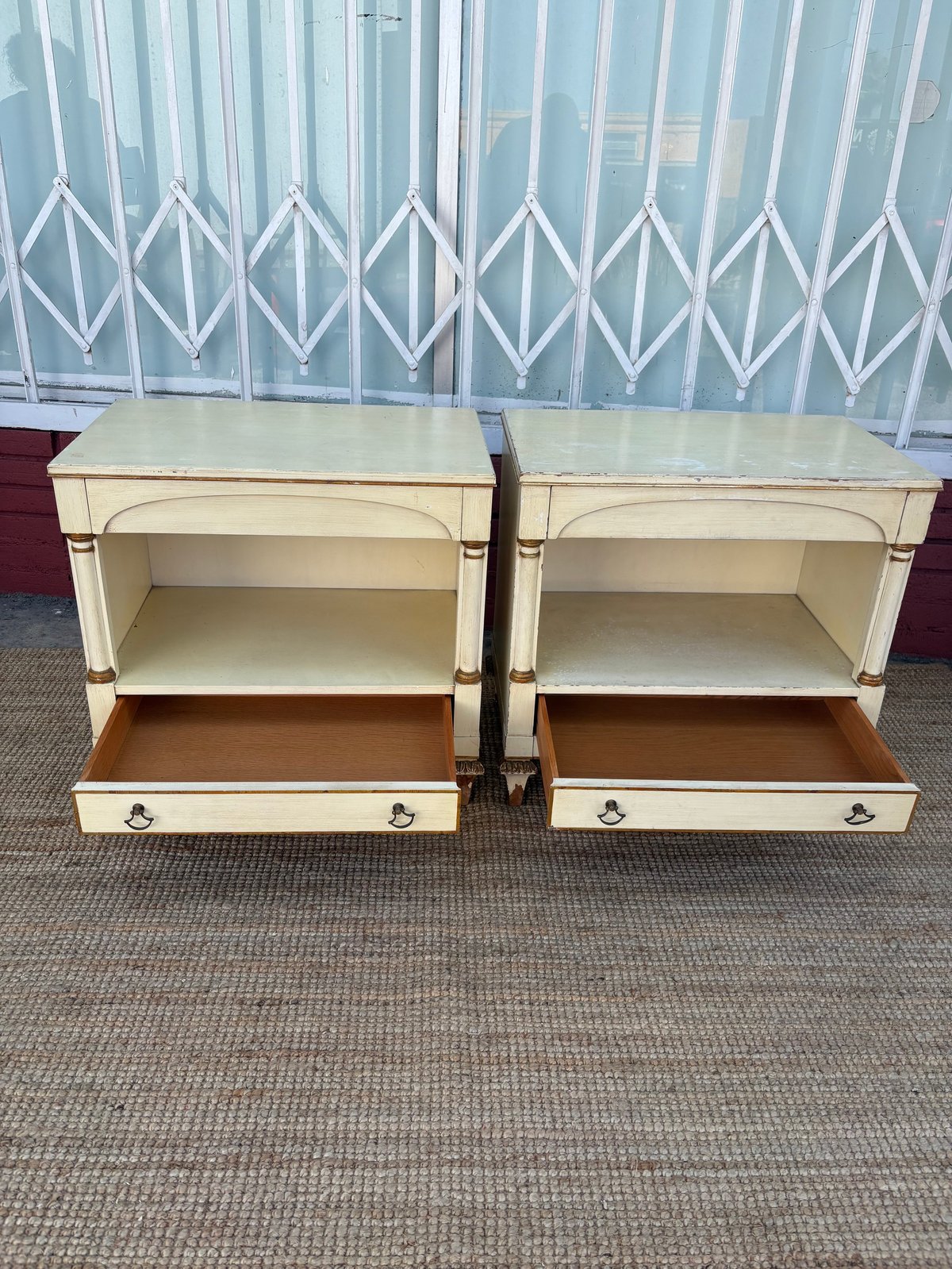 Two cream-colored wooden nightstands with open shelving and bottom drawers against a sheer curtained window