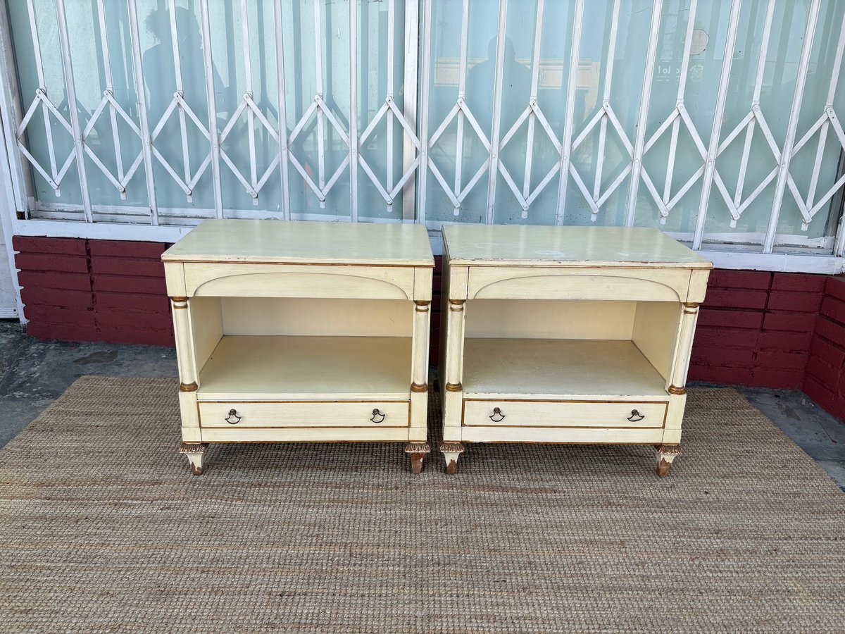 Two cream-colored wooden nightstands with open shelves and drawers on a gray rug against a corrugated metal backdrop