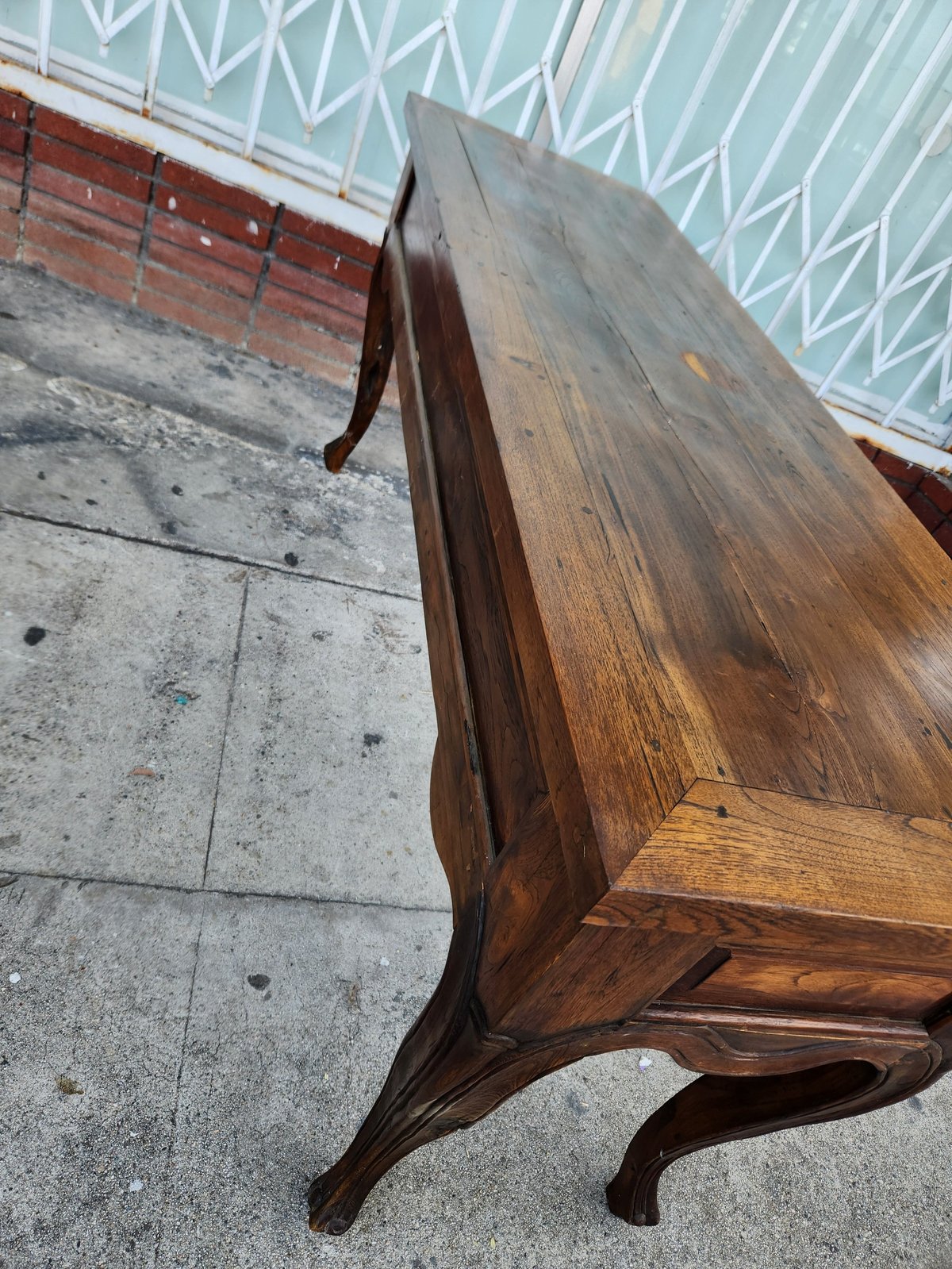 Wooden bench with curved armrests sitting on concrete ground against a metal fence and shed wall