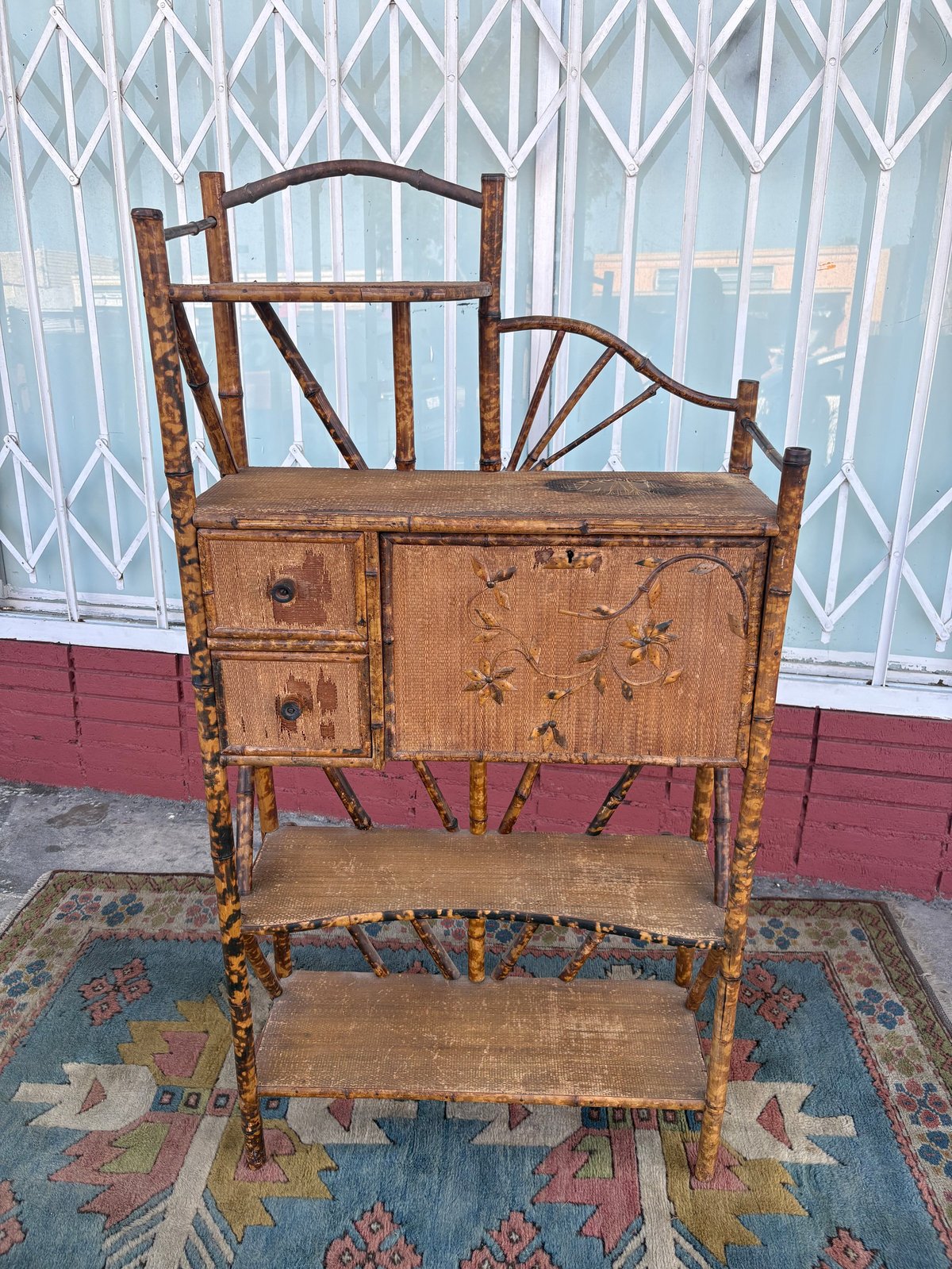 Antique wooden furniture piece with decorative back and storage compartment, placed on a colorful rug against a white and red metal gate