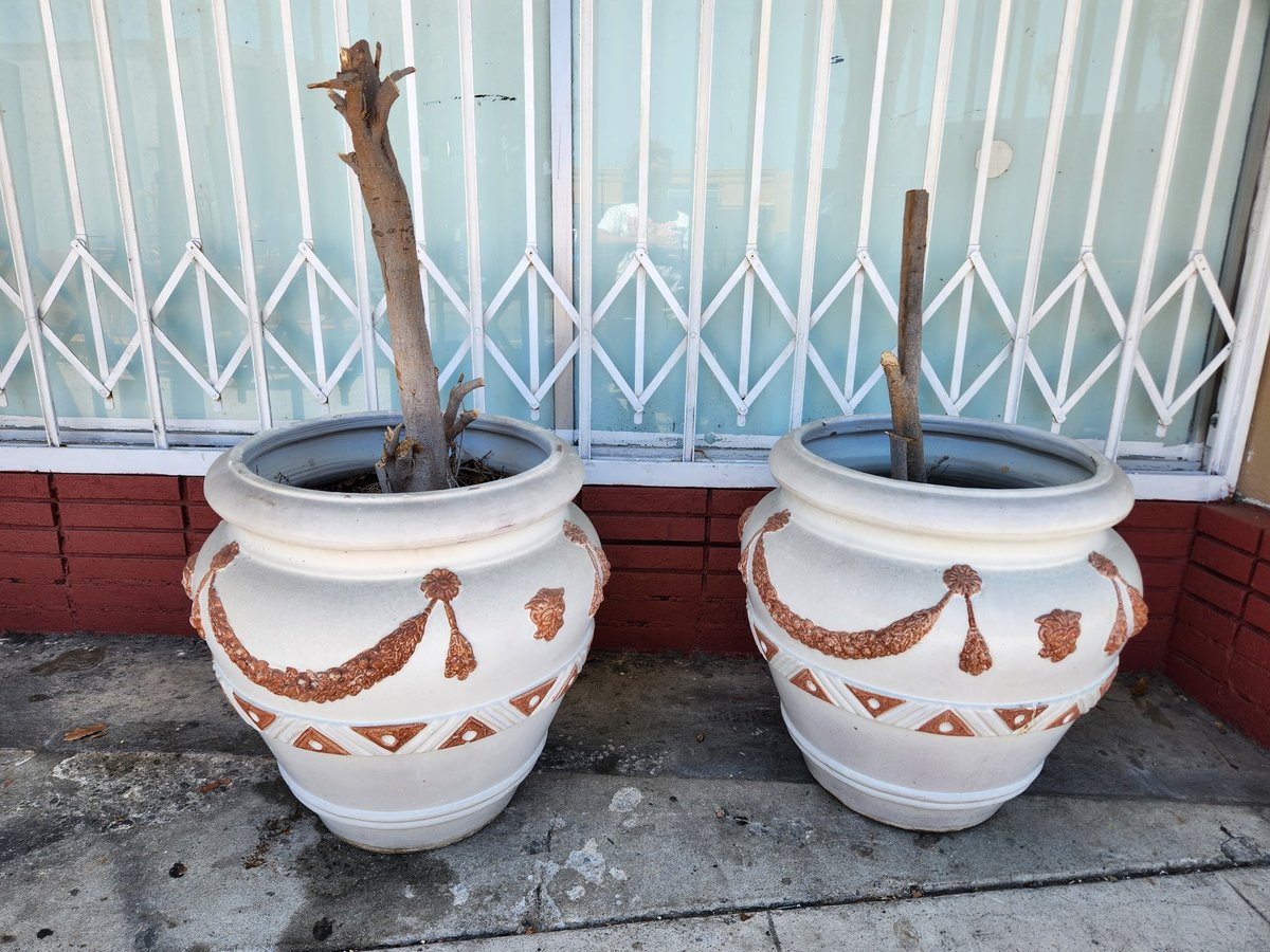 Two white decorative planters with brown geometric patterns containing bare branches, set on pavement against a white metal gate
