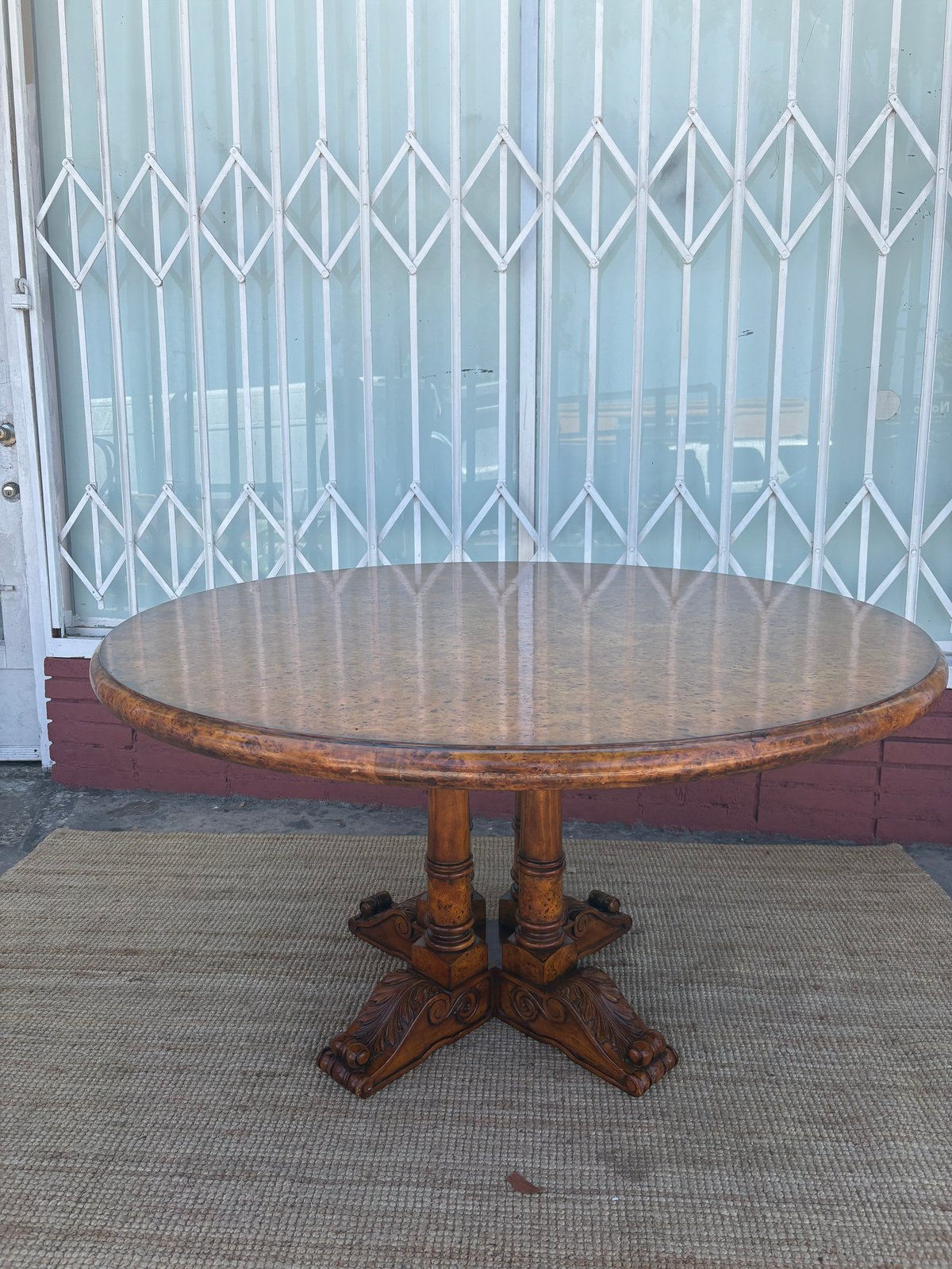 Wooden round pedestal table with ornate base displayed against a metal accordion gate and red brick wall