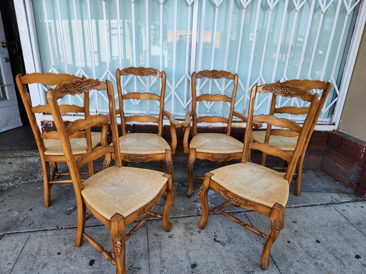 Six wooden dining chairs with woven rush seats arranged outdoors against a corrugated metal wall