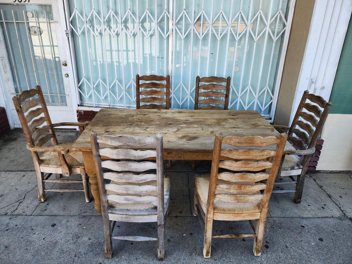 Weathered wooden dining table with six mismatched ladder-back chairs arranged outdoors against a blue curtained backdrop