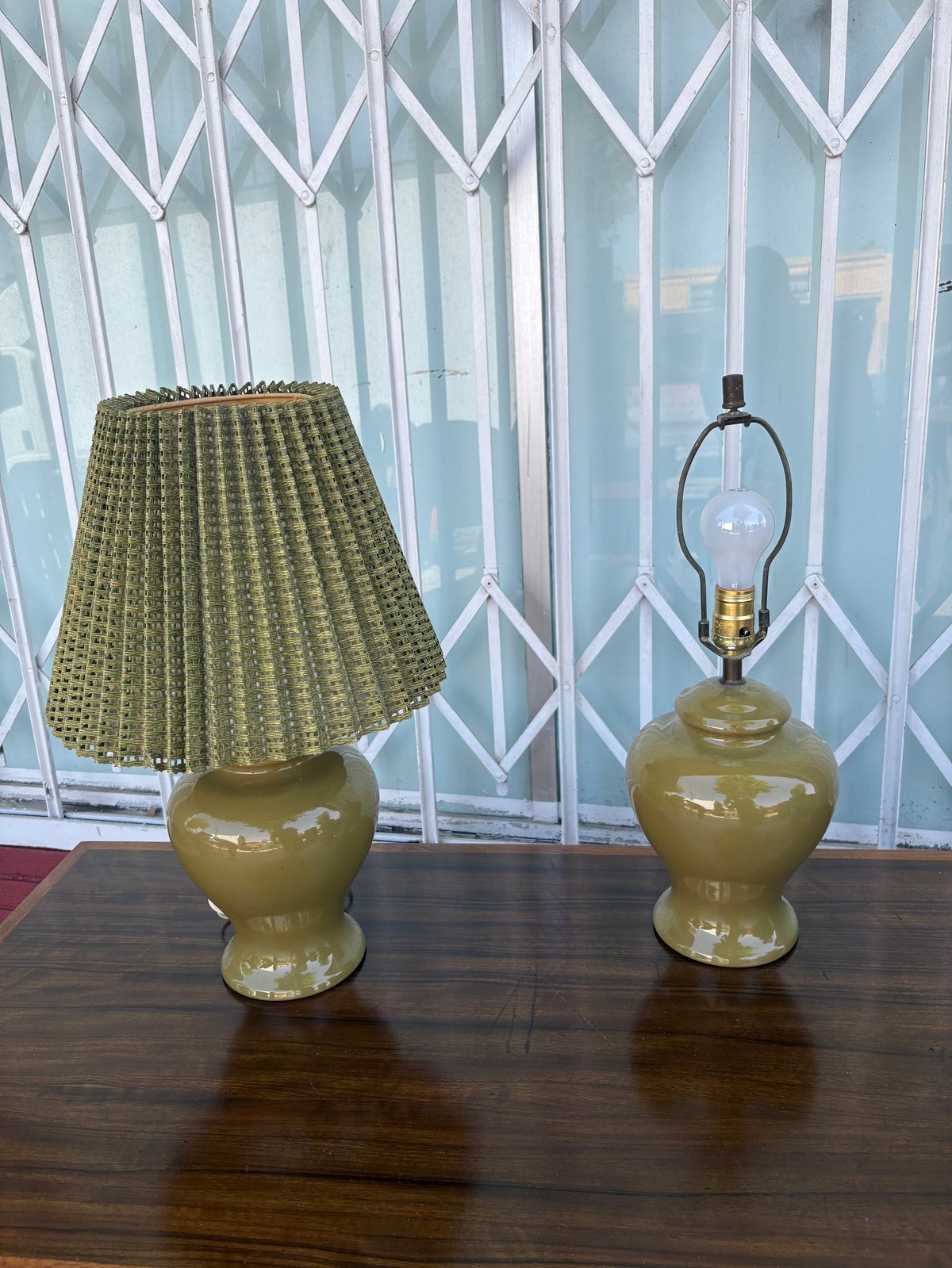 Two olive green ceramic table lamps with pleated fabric shade on wooden surface in front of geometric patterned curtains