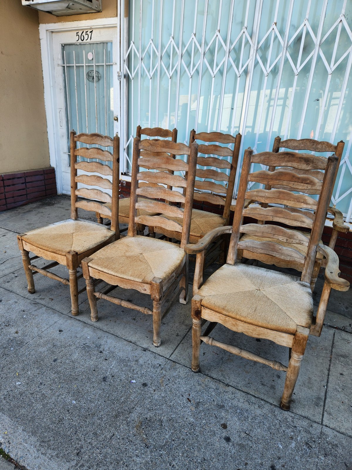 Four wooden rustic chairs with woven rush seats arranged on a concrete patio outside a house with white metal doors