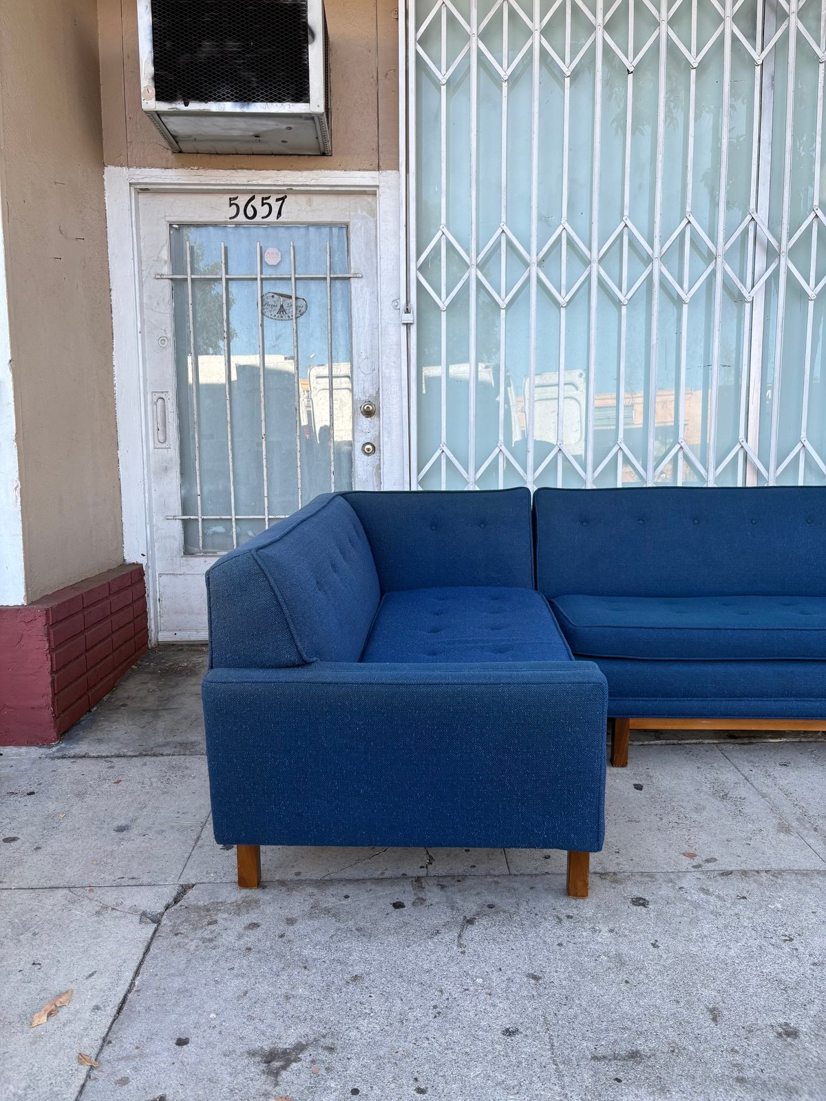 Blue sectional sofa on a sidewalk in front of a building, metal security gate, and air conditioning unit