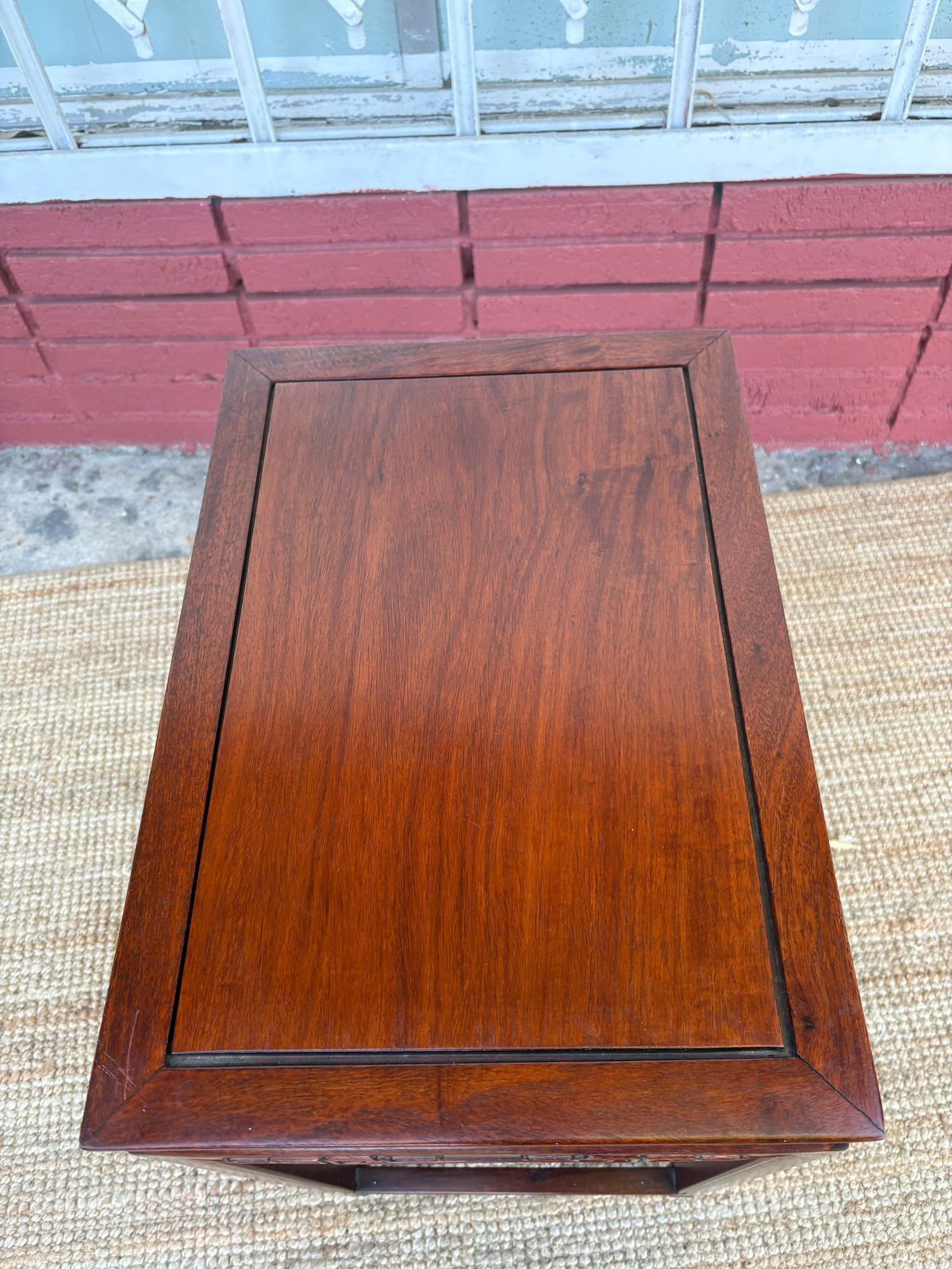 Brown wooden side table with raised frame panel on woven rug against red and blue exterior wall