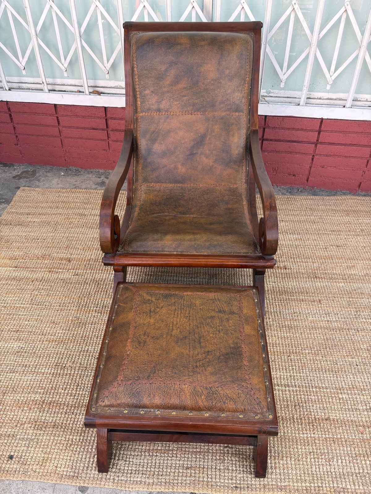 Vintage wooden lounge chair with brown leather seat and matching ottoman on outdoor patio with geometric patterned fence background